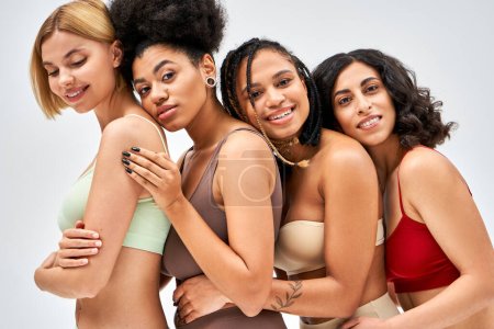 Portrait of cheerful multiethnic women in colorful lingerie hugging each other and posing while standing isolated on grey, different body types and self-acceptance concept, multicultural models