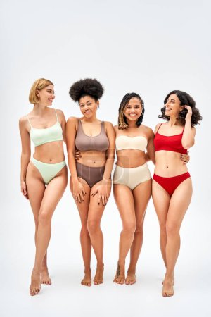Volle Länge positiver und barfüßiger multiethnischer Frauen in bunten Dessous, die sich umarmen und auf grauem Hintergrund stehen, verschiedene Körpertypen und Selbstakzeptanzkonzepte, multikulturelle Modelle