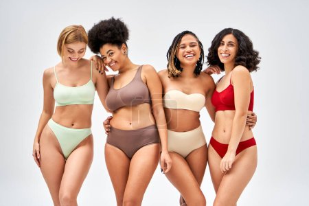 Multiethnic women in colorful lingerie hugging each other and looking at camera while standing isolated on grey, different body types and self-acceptance concept, multicultural models