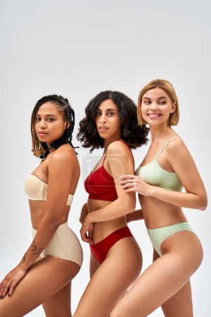 Smiling blonde woman in lingerie looking away while standing near multiethnic friends and posing isolated on grey, different body types and self-acceptance concept, multicultural models