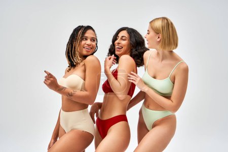 Laughing multiethnic models in modern and colorful lingerie talking while posing and standing together isolated on grey, different body types and self-acceptance concept, multicultural models