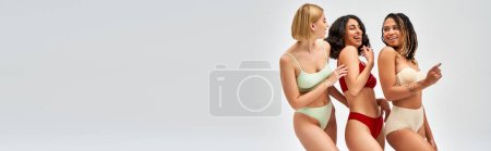 Laughing multiethnic women in colorful and trendy lingerie talking while posing and standing isolated on grey, different body types and self-acceptance concept, multicultural models, banner 