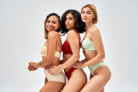 Smiling and multiethnic women in stylish and colorful lingerie hugging and posing while looking at camera isolated on grey, different body types and self-acceptance concept, multicultural models