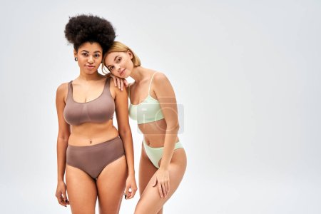 Young blonde woman in trendy lingerie hugging african american friend and looking at camera isolated on grey, different body types and self-acceptance concept, multicultural models