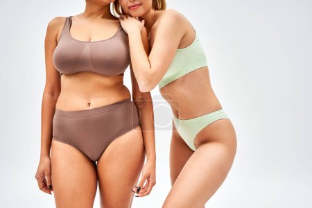 Cropped view of young woman in trendy lingerie hugging african american friend and standing together isolated on grey, different body types and self-acceptance concept, multicultural models