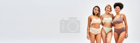 Blonde Frau in moderner Dessous, die afrikanische amerikanische Freunde umarmt und isoliert auf graue Kamera blickt, verschiedene Körpertypen und Selbstakzeptanzkonzept, multikulturelle Models, Banner 