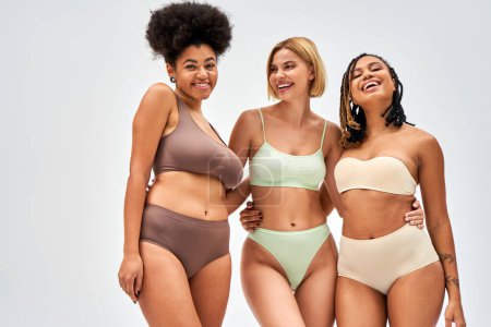 Laughing blonde woman in modern lingerie hugging african american friends while posing together isolated on grey, different body types and self-acceptance concept, multicultural models