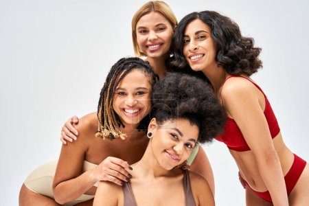 Mujeres multiétnicas positivas en lencería colorida y moderna mirando a la cámara mientras posan juntas aisladas en gris, diferentes tipos de cuerpo y concepto de autoaceptación, modelos multiculturales