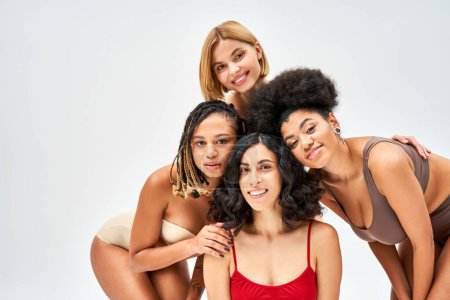 Positive multiethnic group of women in colorful and stylish lingerie hugging and posing together isolated on grey, different body types and self-acceptance concept, multicultural models