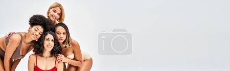 Photo for Joyful and multiethnic women in colorful and trendy lingerie hugging and looking at camera while posing isolated on grey, different body types and self-acceptance concept, multicultural models, banner - Royalty Free Image