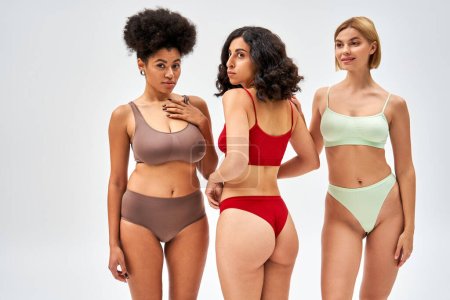 Smiling blonde woman in modern lingerie standing next to multicultural friends in bras and panties on grey background, diverse body shapes and multiethnic women concept