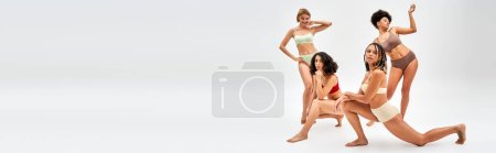 Young african american woman in modern bra and panties posing and standing near multiethnic girlfriends on grey background, different body types and self-acceptance concept with copy space, banner