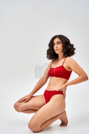 Full length of sexy middle eastern brunette woman in red lingerie holding hand on hip and looking at camera while posing on grey background, self-acceptance and body positive concept 