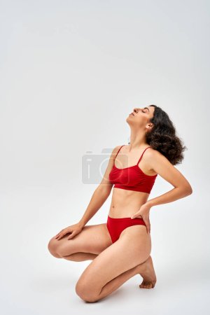 Téléchargez les photos : Vue latérale de la femme brune du Moyen-Orient en lingerie rouge moderne tenant la main sur la hanche et posant avec les yeux fermés sur fond gris, acceptation de soi et concept positif du corps - en image libre de droit
