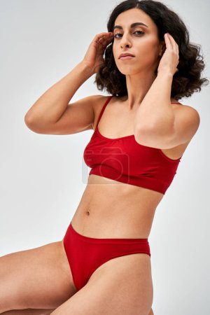 Confident and middle eastern brunette woman in modern red panties and bra touching hair and looking at camera while posing isolated on grey, body acceptance concept 