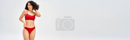 Foto de Mujer positiva de Oriente Medio en sujetador rojo y bragas tocando el cabello y mirando hacia otro lado mientras está de pie y posando aislado en gris, concepto de aceptación del cuerpo con espacio de copia, pancarta - Imagen libre de derechos