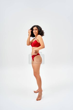 Full length of barefoot and middle eastern woman in modern red lingerie touching hair and looking at camera while standing on grey background, self-acceptance and body positive concept 