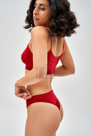 Téléchargez les photos : Femme brune confiante et du Moyen-Orient en soutien-gorge rouge touchant culotte et détournant les yeux tout en se tenant debout et posant isolé sur gris, l'acceptation de soi et le corps concept positif - en image libre de droit