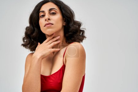 Portrait de femme brune du Moyen-Orient en soutien-gorge rouge tendance touchant le cou et regardant la caméra tout en étant isolé sur le gris, l'acceptation de soi et le concept positif du corps 
