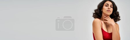 Téléchargez les photos : Femme brune confiante du Moyen-Orient en soutien-gorge rouge moderne touchant le cou et regardant la caméra tout en se tenant debout et posant isolée sur le concept gris, l'acceptation de soi et le corps positif, bannière - en image libre de droit