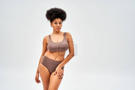 Confident african american woman in modern brown bra and panties looking at camera while touching hip and posing isolated on grey with copy space, self-acceptance and body positive concept 
