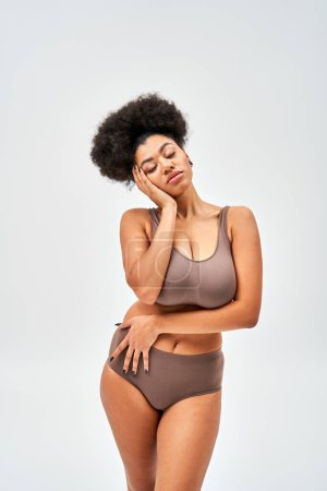 Seductive african american woman in modern brown lingerie touching cheek while posing with closed eyes and standing isolated on grey, self-acceptance and body positive concept 