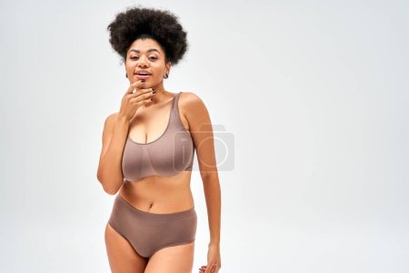 Foto de Mujer afroamericana positiva y seductora en bragas marrones y sujetador tocando la barbilla y mirando a la cámara mientras está de pie aislado en gris, auto-aceptación y el concepto de cuerpo positivo - Imagen libre de derechos