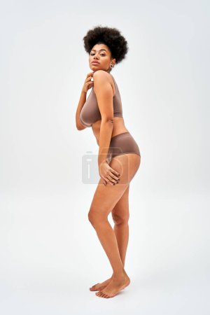 Full length of barefoot and sexy african american model in modern brown lingerie touching shoulder and looking at camera on grey background, self-acceptance and body positive concept 