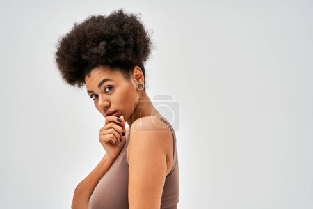 Portrait of confident african american woman in modern brown bra touching chin and looking at camera while standing isolated on grey, self-acceptance and body positive concept 