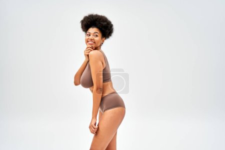 Modèle afro-américain joyeux en lingerie marron touchant le menton et détournant les yeux tout en se tenant debout et posant isolé sur un concept gris, d'acceptation de soi et de corps positif 
