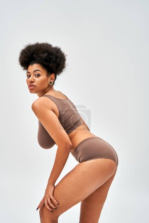 Confident and modern african american woman in brown lingerie looking at camera while touching leg and posing isolated on grey, self-acceptance and body positive concept 