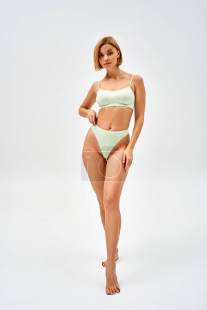Photo for Full length of confident young blonde woman in light green lingerie looking at camera while standing and posing on grey background, self-acceptance and body positive concept - Royalty Free Image