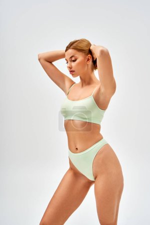 Sensual young woman with natural makeup wearing light green lingerie and touching blonde hair while looking down and standing isolated on grey, self-acceptance and body positive concept 