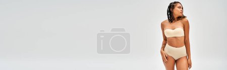 Foto de Moderno modelo afroamericano joven con maquillaje natural usando lencería beige sexy y tocando la cadera aislada en gris, autoaceptación y concepto positivo para el cuerpo, pancarta con espacio para copiar - Imagen libre de derechos