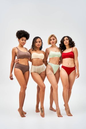 Volle Länge von sexy und barfuß multiethnischen Frauen in bunten Dessous, die sich umarmen und in die Kamera schauen auf grauem Hintergrund, multikulturelle Models und Body Positivity Bewegungskonzept