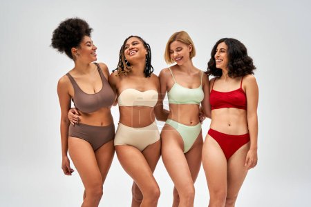 Positive and multiethnic confident women in colorful lingerie hugging while looking at each other and standing isolated on grey, multicultural models and body positivity movement concept