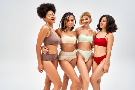Cheerful and modern multiethnic girlfriends in colorful lingerie hugging each other while posing isolated on grey, multicultural models and body positivity movement concept