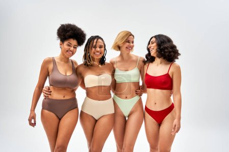 Cheerful multiethnic girlfriends with natural makeup hugging while posing in colorful lingerie and standing isolated on grey, multicultural models and body positivity movement concept