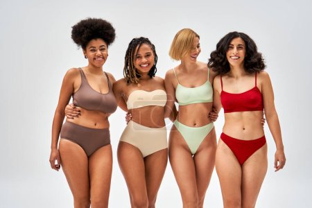 Fröhliche und sexy multiethnische Frauen in bunten Dessous, die sich umarmen und gemeinsam in die Kamera schauen, isoliert auf grauen, unterschiedlichen Körpertypen und Selbstakzeptanz, multikulturelle Repräsentation