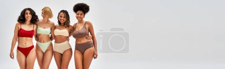 Laughing and confident multiethnic women in colorful lingerie hugging and posing together isolated on grey, different body types and self-acceptance, multicultural representation, banner 