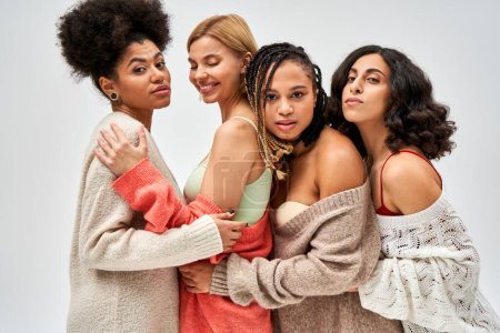 Portrait of group of multiethnic girlfriends in warm knitted jumpers hugging and standing together isolated on grey, different body types and self-acceptance, multicultural representation