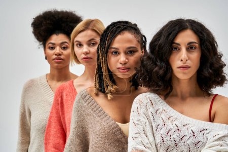 Retrato de mujeres multiétnicas en suéteres cálidos elegantes mirando a la cámara cerca de demonios borrosos aislados en gris, diferentes tipos de cuerpo y la auto-aceptación, la representación multicultural