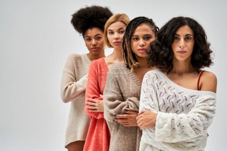 Selbstbewusste und multiethnische Frauen in stylischen Strickpullovern, die sich die Arme verschränken und nebeneinander stehen, isoliert auf grauen, unterschiedlichen Körpertypen und Selbstakzeptanz, multikulturelle Repräsentation
