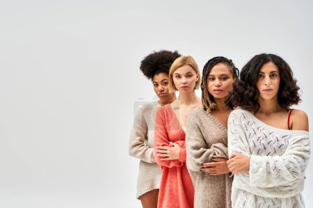 Grupo multiétnico de mujeres en suéteres de punto elegantes cruzando los brazos y mirando a la cámara aislada en gris, diferentes tipos de cuerpo y auto-aceptación, representación multicultural, espacio de copia