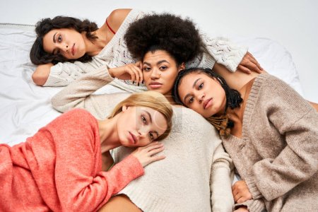 High angle view of trendy and multiethnic women in warm and knitted jumpers lying together on bed isolated on grey, different body types and self-acceptance, multicultural representation