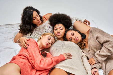 High angle view of multiethnic women in stylish warm jumpers looking at camera while lying on bed isolated on grey, different body types and self-acceptance, multicultural representation