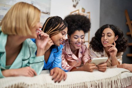Positive afrikanisch-amerikanische Frau hält Smartphone in der Nähe verschwommener multiethnischer Freundinnen im bunten Pyjama auf dem Bett liegend während Pyjama-Party im Schlafzimmer zu Hause, Bindungszeit in bequemer Nachtwäsche