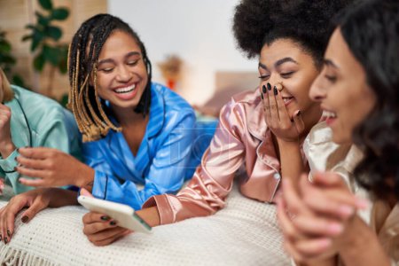 Téléchargez les photos : Femme afro-américaine gaie couvrant la bouche et utilisant un smartphone tout en s'amusant avec des copines multiethniques en pyjama coloré sur le lit à la maison, liant le temps en vêtements de nuit confortables - en image libre de droit