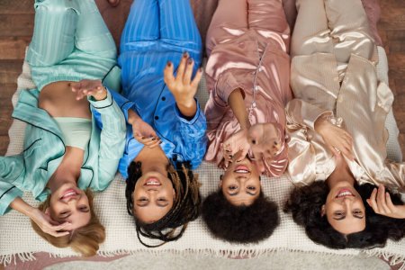 Vista superior de las novias sonrientes y multiétnicas en pijama colorido extendiendo las manos y mirando a la cámara durante la fiesta de pijama en la cama en el dormitorio en casa, tiempo de unión en ropa de dormir cómoda