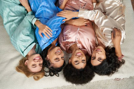 Vista superior de alegres novias multiétnicas en pijama colorido abrazándose y mirando a la cámara mientras están acostados en la cama durante la fiesta de pijama en casa, tiempo de vinculación, fiesta de pijamas